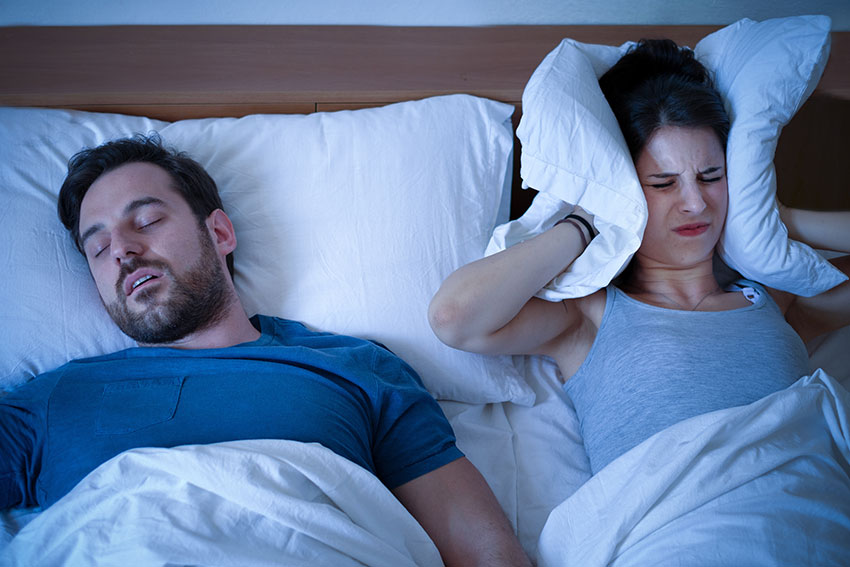 woman holding a pillow tightly to her ears while husband snores loudly next to her in bed